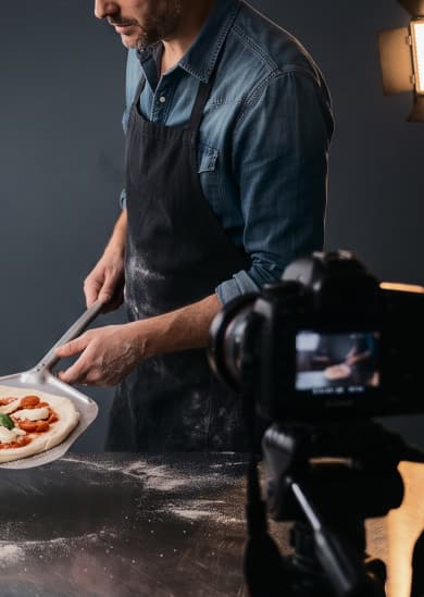 An instructor in an apron slides a topped pizza onto a metal peel at a stainless-steel counter, with a video camera and studio lights beside them