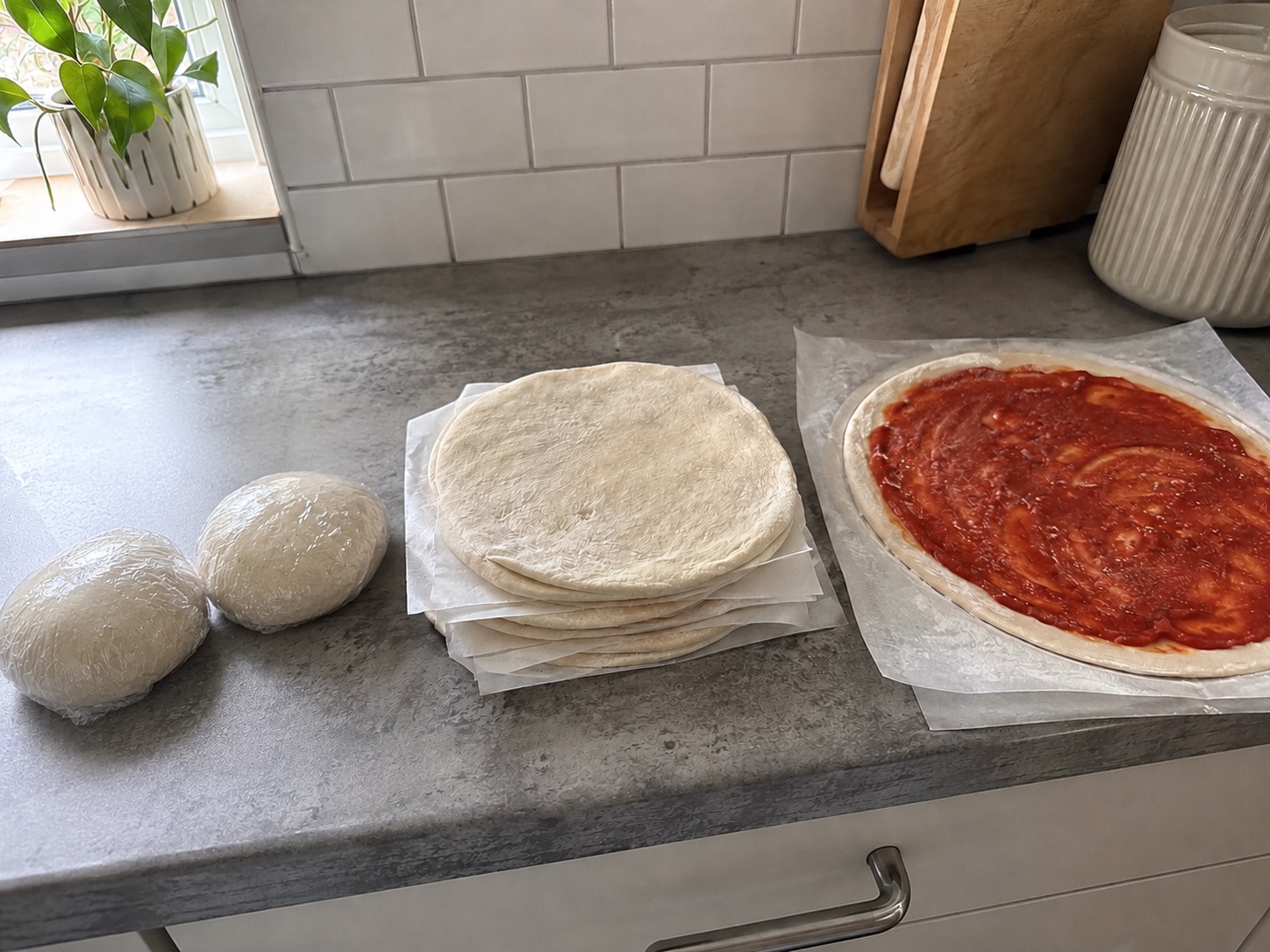 Tre modi per congelare l'impasto della pizza sul bancone della cucina: due palline avvolte nella pellicola, una pila di dischi stesi separati da carta forno e una base condita con salsa di pomodoro
